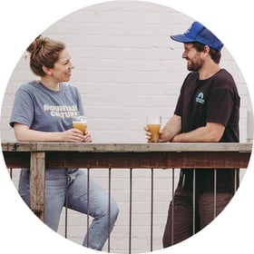 founders of mountain culture harriet and dj mccready facing each other holding each glass pint of beer leaning over outdoor railings