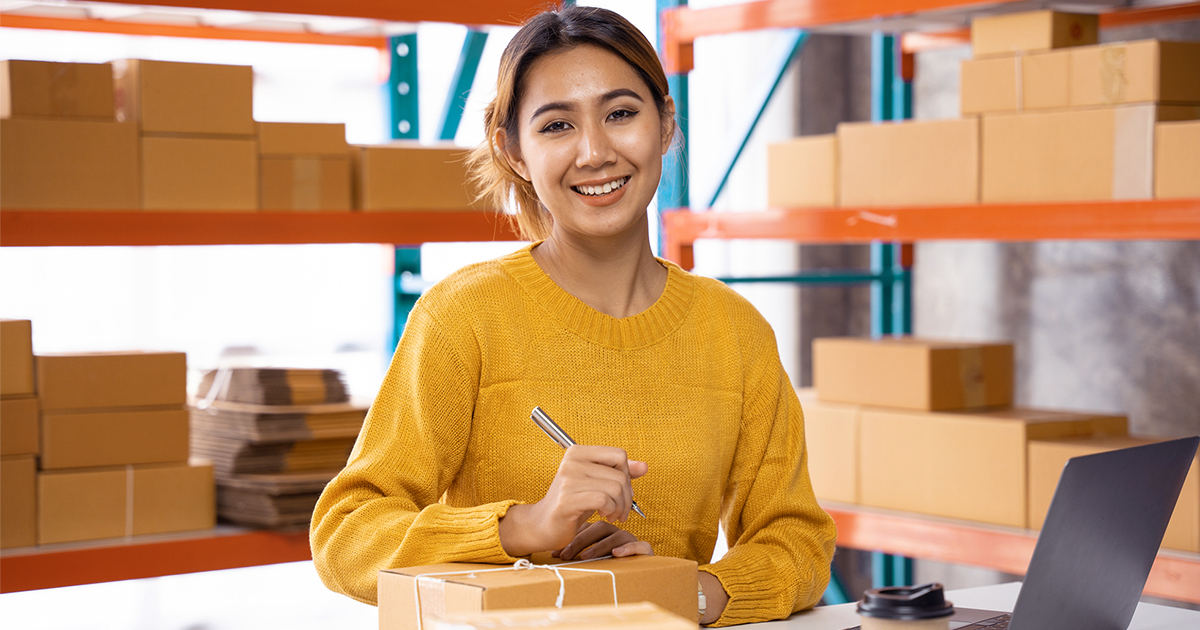 woman in shipping warehouse package shelves behind working on desk with packages and laptop