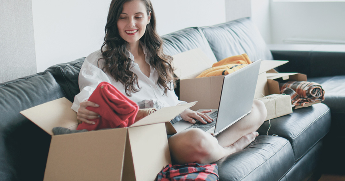 woman sitting on couch holding laptop in between boxes of clothes