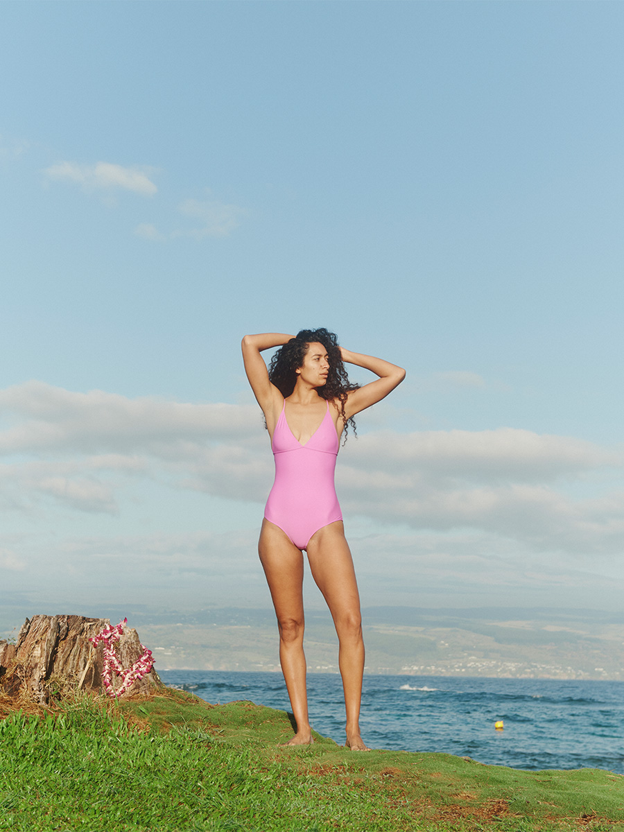 woman wearing pink pali swim swimwear standing near cliff side overlooking sea tree stump at the corner