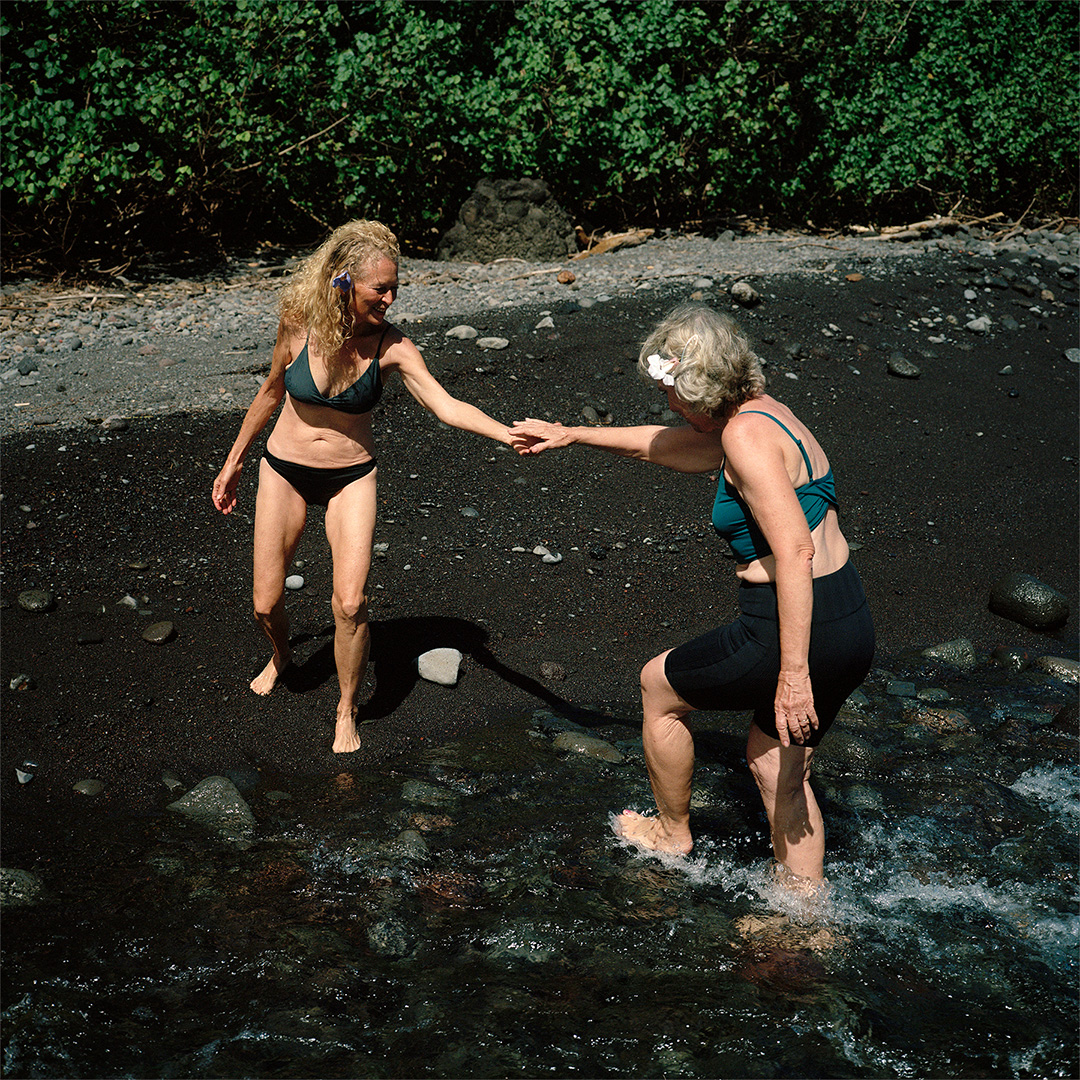 two happy women holding hands reaching each other along the river banks wearing pali swim swimwear