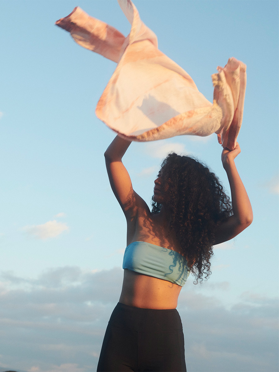 woman wearing pali swim swimwear standing raised arms swinging linen top upward in the air sunset sky in the background