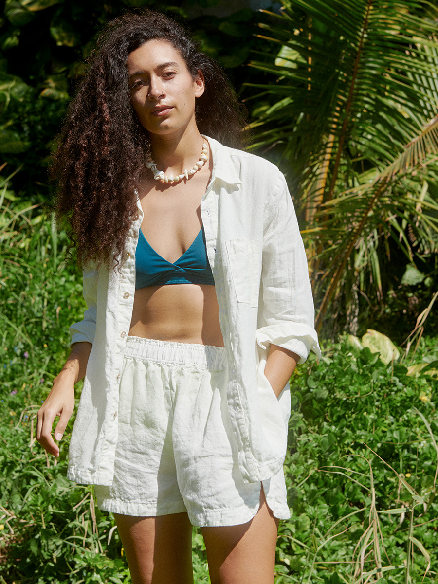 woman wearing pali swim swimwear and linen top and bottoms standing in the sun in front of foliage in the background