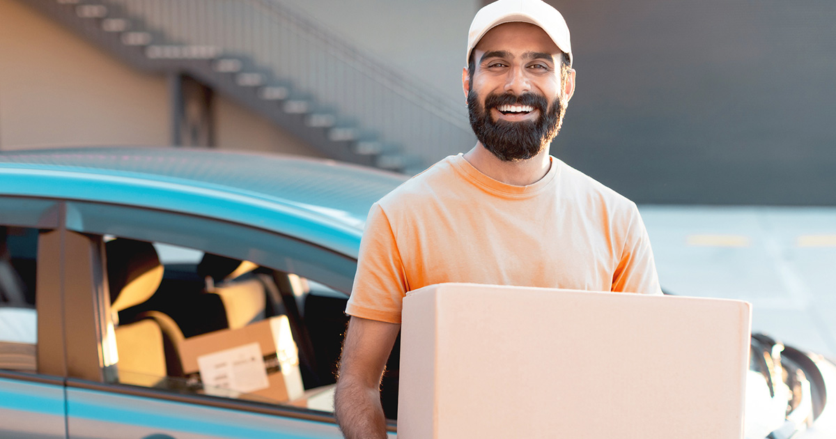 happy indian delivery man standing near car holding carton box parcel