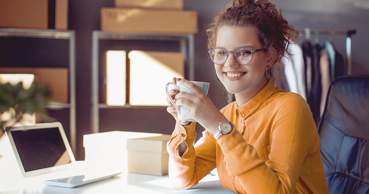 woman sitting at the desk holding mug laptop boxes in front boxes shelves behind